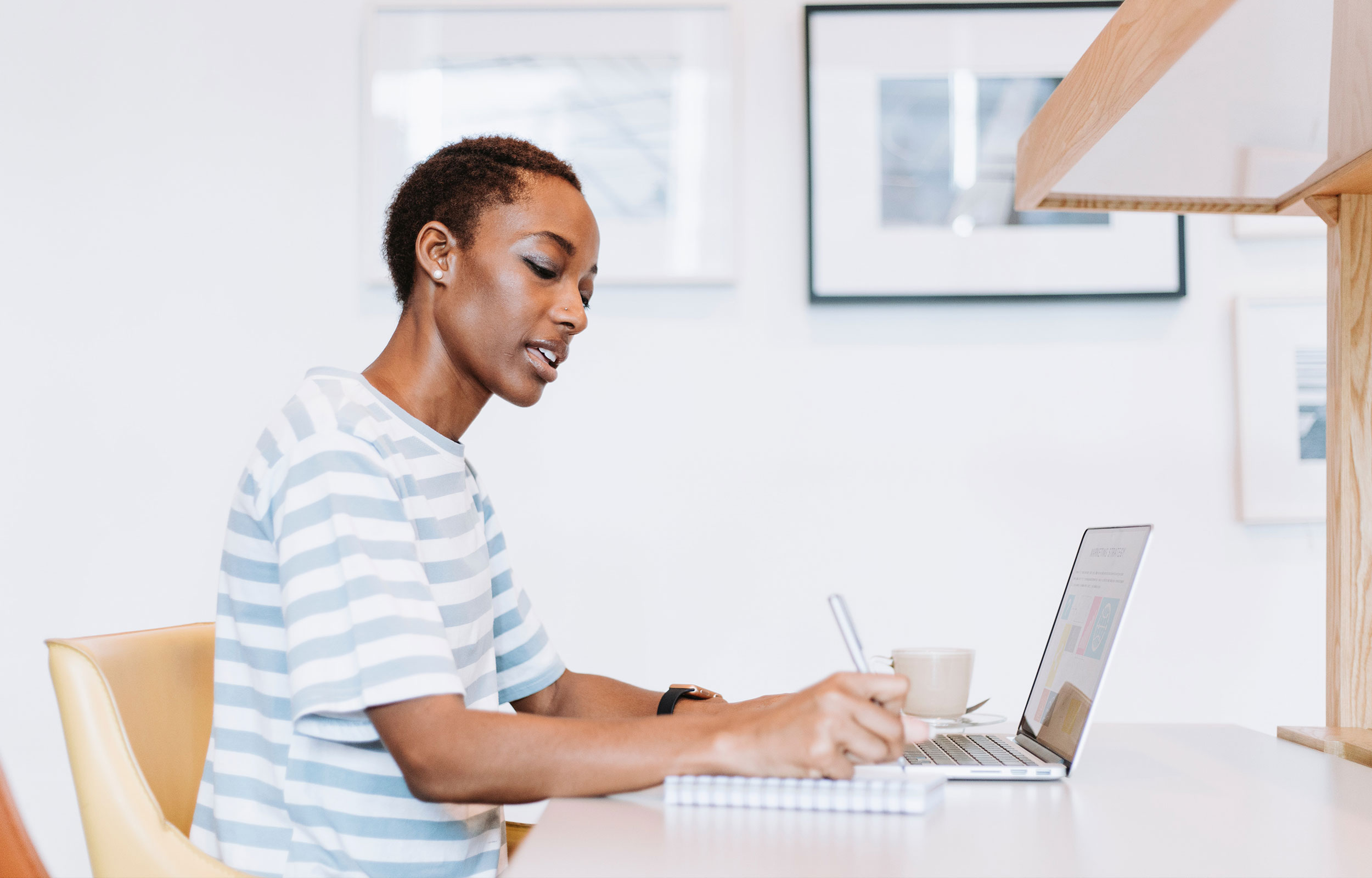 Person writing in a notebook while working on a laptop at a bright desk.