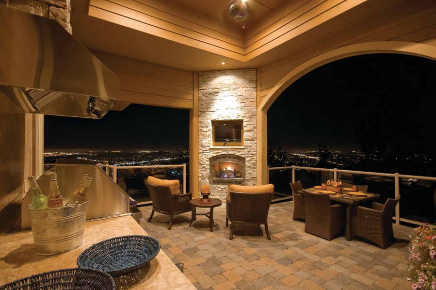 An illuminated patio at night with several chairs, a fireplace, and outdoor television. The deck looks over the city at night-time.